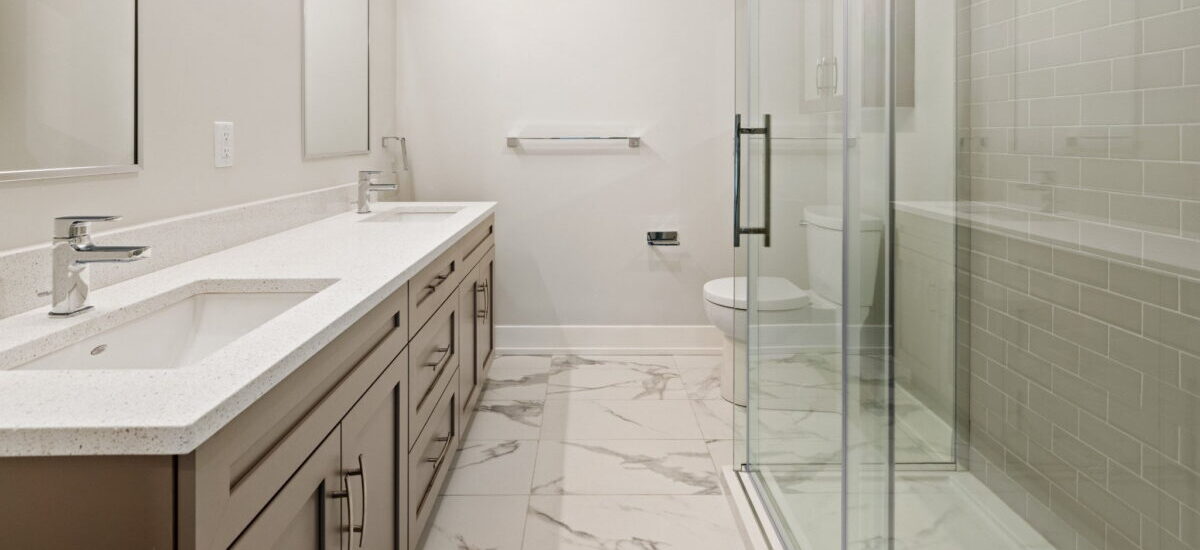 A bathroom with a two sink counter on the left. The counter is white and the cabinets beneath are medium brown. On the right a glass encased shower with grey subway tile is seen, with a toiler just visible beyond it.