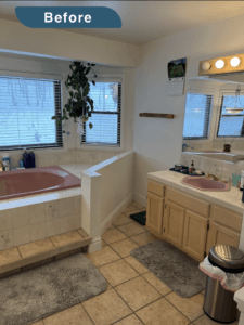 An image labeled "Before." A bathroom with pastel pink ceramic tub and sink. The wood cabinets are a blonde finish. The tile floor is made of large square tiles that are a tan spattered with brown style. There are two large gray rugs. Mini-blinds cover the window.s