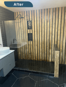An image labeled "After." A gorgeous shower with wood slat walls and black tiled shower floor. It is open with a glass panel separating it from the rest of the bathroom. There are two black tile cutouts in the wood slats to hold toiletries. The basin sink is set on a white countertop with grey cabinetry underneath.